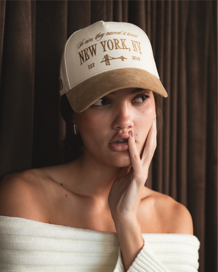Woman wearing a beige and brown cap with 'New York, NY' text against a brown curtain background