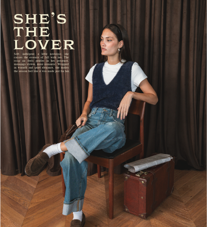 Woman sitting on a chair with a suitcase next to her, wearing a blue vest and jeans, with text 'SHE'S THE LOVER' above.