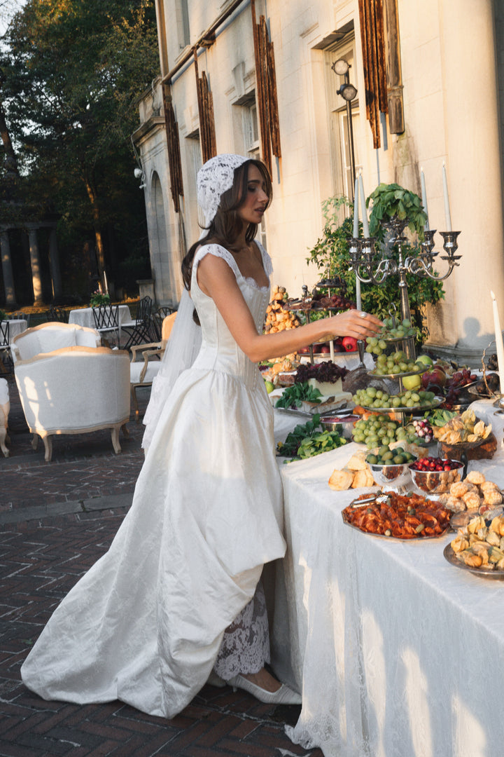 Danielle Bernstein in a white dress standing by a table with food outdoors.