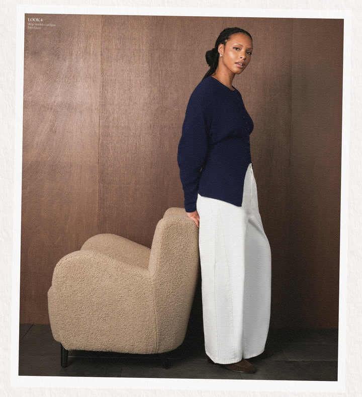 Woman in a navy sweater and white pants standing next to a beige armchair against a brown background