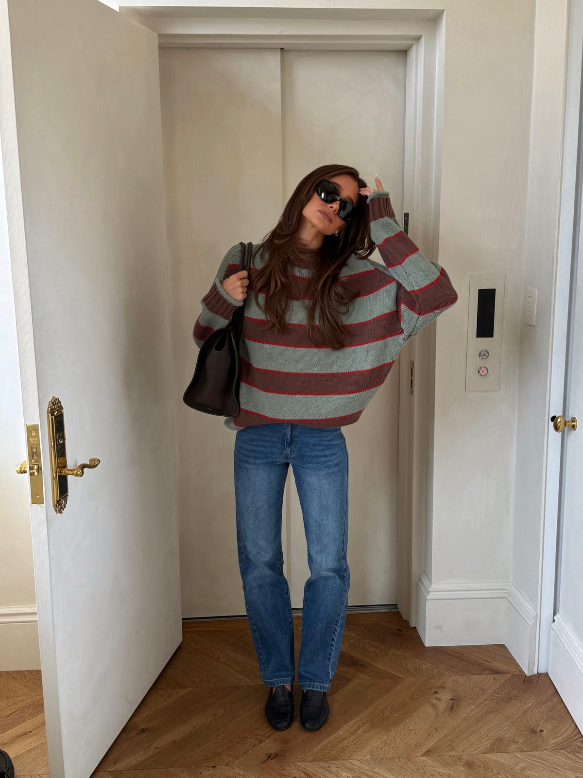 A woman wearing a muted green and brown striped knit sweater, blue jeans, and black shoes stands in a doorway holding a black shoulder bag.