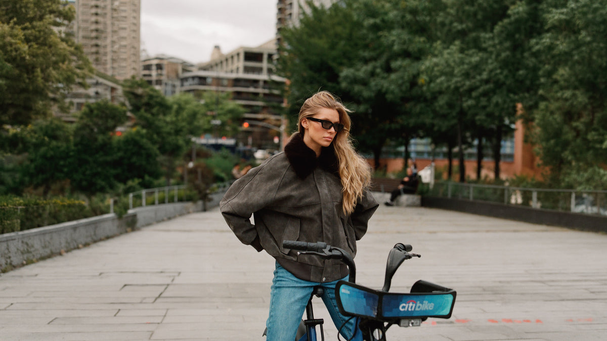 A woman wearing a dark grey fur-trimmed patina bomber jacket and blue jeans stands on a Citibike in an urban park setting.