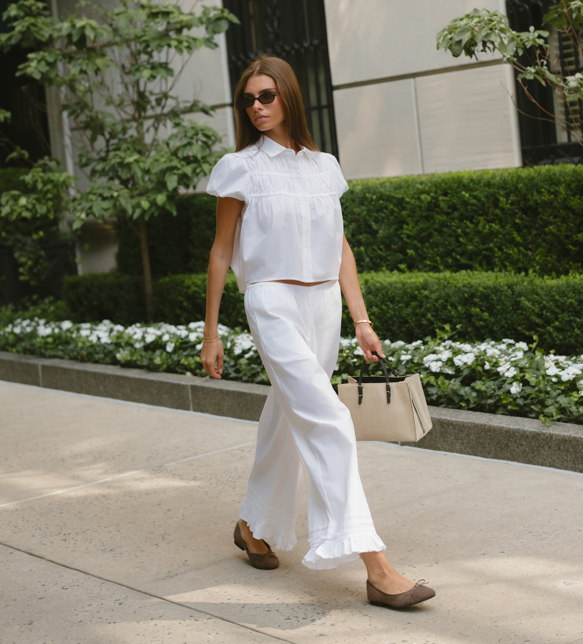 A woman walking outdoors on a sidewalk wearing a white short-sleeve cropped blouse and matching white wide-leg pants, paired with taupe flats and carrying a beige handbag.